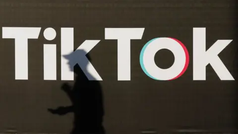 Getty Images A young man holding a smartphone casts a shadow as he walks past an advertisement for social media company TikTok.