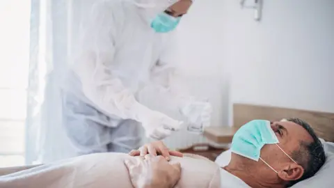 Getty Images man in mask in hospital bed with medic in full PPE