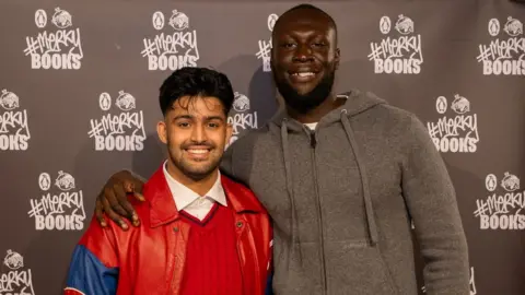 #Merky Books  Sufiyaan Salam pictured with Stormzy after he won the New Writers' Prize. Sufiyaan is a 26-year-old British Asian man with his dark hair long on top but shaved close on the sides. He has brown eyes and a short beard and moustache. He wears a red leather jacket over a red jumper and white shirt. Stormzy stands about a foot taller to his left. The rapper is a black man in his 30s with cropped hair and a short beard. He has his arm around Sufiyaan's shoulders and wears a grey hoodie. They're photographed in front of #Merky Books event hoarding