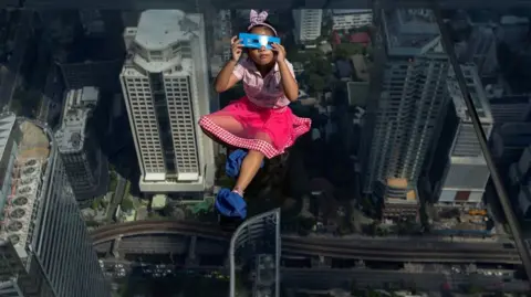 A girl looks through an eclipse viewer on an elevated, transparent platform, with buildings and roads in the distance below her