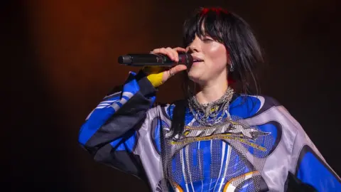 Getty Images Billie Eilish performing on stage in August 2023. Billie is a 22-year-old white woman with long black hair worn loose. She has a fringe to her eyebrows and wears a graphic long-sleeved blue and grey top. She holds a microphone up as she sings.