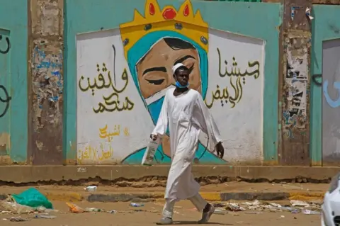AFP A man walks by a mural. It features a woman's face wearing a mask. There is some litter on the street.