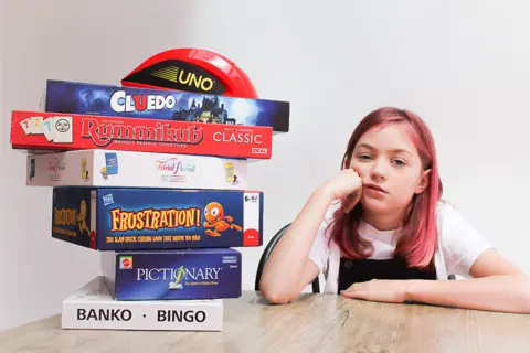  Emily Pritchett  A girl looks bored next to a stacked pile of board games