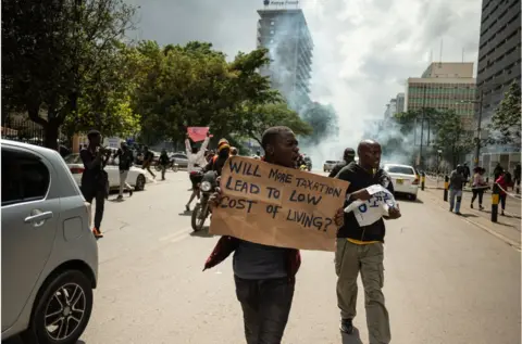 Edwin Ndeke/ Getty Images Protesters walking through the street, one carrying a placard which reads: Will more taxation lead to low cost of living.