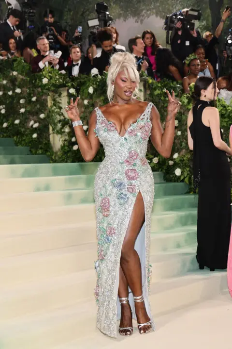 JUSTIN LANE/EPA Aya Nakamura wears an ivory dress encrusted in pearlescent flowers. Her wig and eyebrows are dyed a vivid shade of blonde.