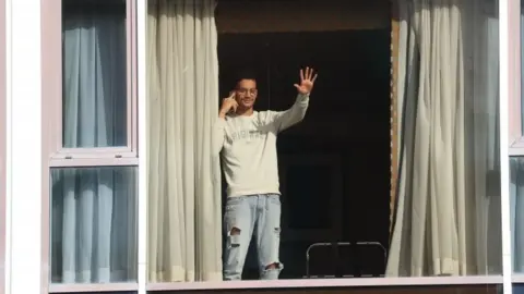 PA Media Roger Goncalves, who is from Brazil and travelled via Madrid to get to the UK, waves to members of the media from the window of Radisson Blu Edwardian Hotel, near Heathrow Airport, London