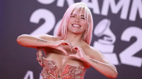 Getty Images Karol G doing a heart sign with her hands, with pink hair and a pink sparkly dress