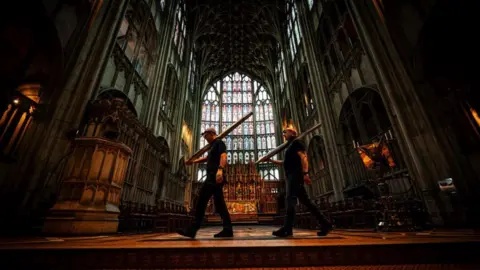 Ben Birchall/PA Wire Two men walking through Gloucester Cathedral. They are both wearing hard hats and carrying equipment.