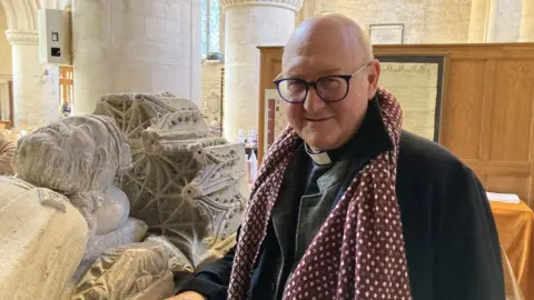 BBC The Reverend Oliver Ross stands in the abbey next to the medieval reconstruction of the empty tomb of King Athelstan.