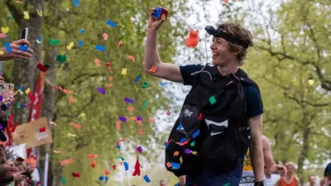 George Scholey running along the course carrying a Rubik's Cube, which he is showing to the crowd