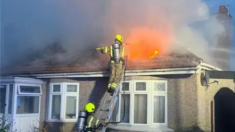 Essex Fire and Rescue Service Fire fighters up a ladder at the front of a bungalow tacking a fire in the roof