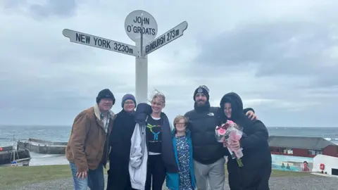 Family photo Iris and her family standing beneath the John O'Groats sign