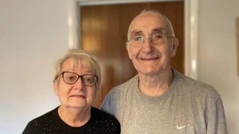 Laura Coffey/BBC A portrait photo of Mary and Allan Bushell standing side by side in their home