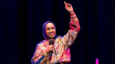 1Xtra Ola Labib performing at BBC 1Xtra Comedy Gala at the Hackney Empire on 1 April 2024. Ola is a British Sudanese woman in her 30s. She has brown eyes and smiles, wearing a blue hijab and a checked-patterned tracksuit in yellow, red and blue. She holds a microphone in her right hand while pointing to the ceiling with her left. The staging behind her is black with pink accents.