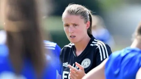 Getty Images Lydia Bedford as Leicester Manager, wearing a black coach's top with white stripes on the shoulder. Players' shoulders are visible in the front of the frame, but indistinct. Lydia's hair is tied back and she looks focused on the action around her.
