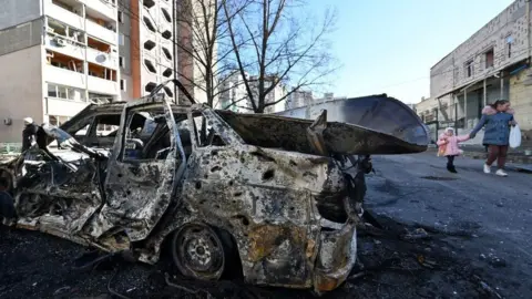 AFP A woman and her child walk past a car destroyed by shelling in the outskirts of Ukraine's capital, Kyiv (28 February 2022)