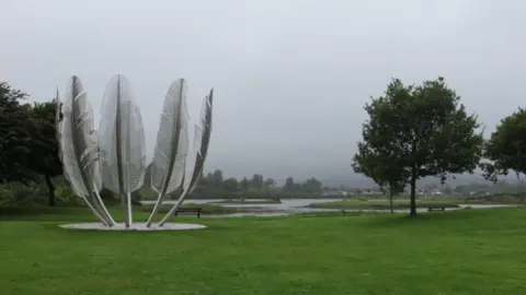 Brian McAleer a stainless steel sculpture of huge eagle feathers in a circle, next to a tree on a cloudy grey day