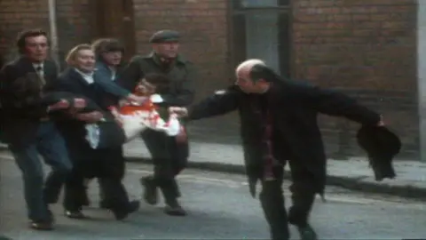 A priest waves a bloodstained handkerchief as four men carry an injured, bloodied man through the streets of Londonderry on Bloody Sunday