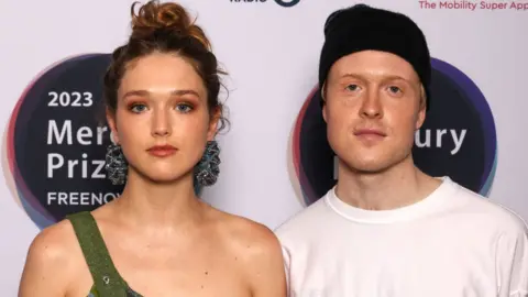 Getty Images Members of Jockstrap, Georgia Ellery and Taylor Skye attend The Mercury Prize 2023 awards show at Eventim Apollo on September 07, 2023 in London. Georgia stands on the left, she wears a green top over one shoulder and large blue hoop earrings. She has blue eyes and her dark curly hair is tied in a bun on her head. She wears brown-orange eyeshadow and a muted red lipstick. Taylor stands on the right, he has blue eyes, freckled skin and fair hair covered by a dark beanie hat. He wears a white T-shirt and both he and Georgia look at the camera with a neutral expression.