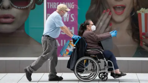 Getty Images A woman sitting in a wheelchair wearing a mask and gloves, being pushed by a man wearing the same