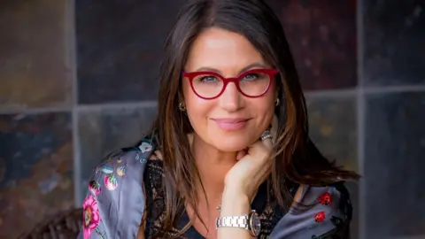 Kat Gebauer Ms Gersh, wearing red glasses and resting her head on one hand, looks at the camera in this portrait