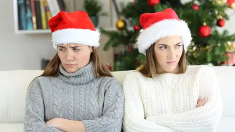 AntonioGuillem Two women wearing Santa hats looking away from each with a Christmas tree in the background