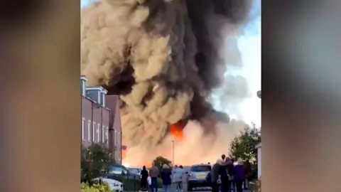 Huge fire with row of buildings in front. People stand and look towards fire.