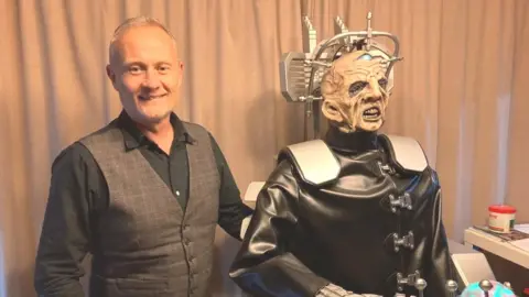 James Grant/BBC Man with very short hair and wearing a waistcoat stands next to dummy of the Davros character