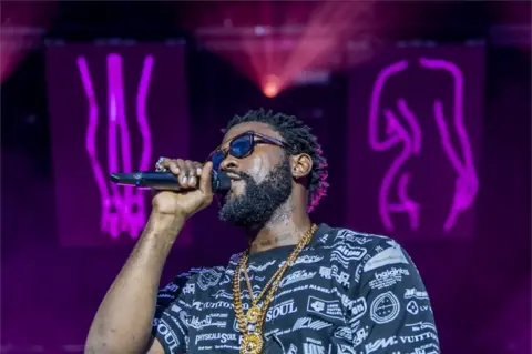 EPA Damso holds the microphone to his mouth as he looks out to the audience. He is wearing sunglasses and a T-shirt covered in logos and behind him are purple neon signs depicting women.