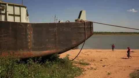 Reuters People walk past a stranded barge on the shore of the Rio Paraguay (Paraguay River), which flows down to the Parana River, as the lack of rain in Brazil, where the river originates, has brought water levels down, forcing cargo ships to reduce the amount of grains that are loaded for export, in Ypane, Paraguay August 30, 2021