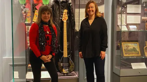 Chelmsford Museum Suzi Quatro and a Chelmsford Museum staff member standing next to the Sterling by Music Man black five-string bass
