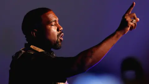 Reuters Kanye West appears to be addressing an audience. He has one arm raised up, his index finger pointing skywards, and looks determined as he speaks. There's a dark blue light emanating from the background, and a faint blue glow can be seen on the underside of his arm where his skin meets the light.