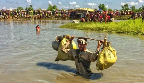 BBC A Rohingya man wading in waist-deep water carries sacks and pots over his head, looking directly at the camera