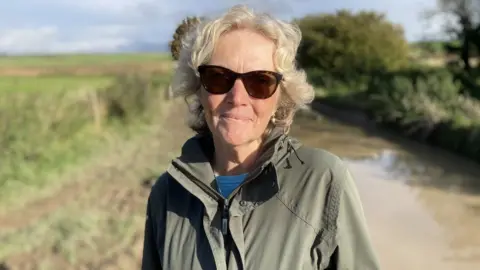 Angela Norman in sunglasses and green raincoat stands in front of the flooded country road, with brown muddy water, green muddy banks and sunshine.