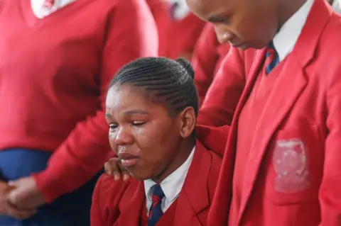 AFP A learner is comforted by another as she weeps while empty coffins used in a symbolic mass memorial service in East London on July 6, 2022, after 21 people, mostly teens, died in unclear circumstances at a township tavern last month, in an incident that shocked South Africa.