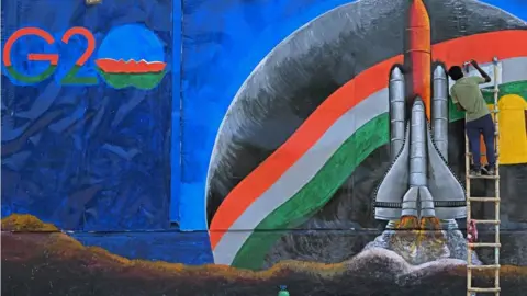 Getty Images TOPSHOT - An artist paints a wall mural surrounding a garbage dump aside a logo of the G20 India summit ahead of its commencement in New Delhi on September 3, 2023. (Photo by Sajjad HUSSAIN / AFP) (Photo by SAJJAD HUSSAIN/AFP via Getty Images)