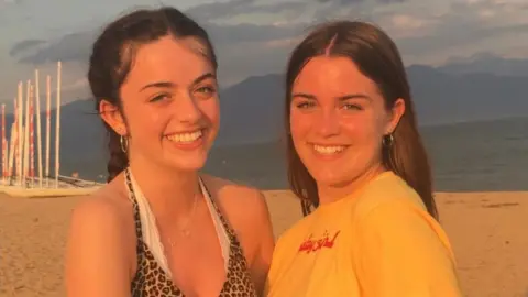 Lawrence family  Two young women with auburn hair, smiling on a beach hugging each other