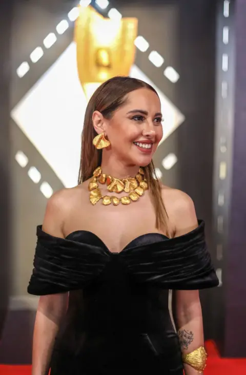 Getty Images Yasmeen Raess dressed in a black dress and gold jewellery smiling on the red carpet.