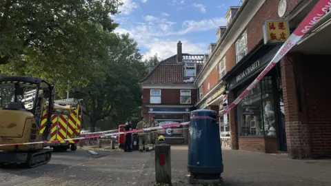 Robby West/BBC Fire and rescue tape across a parade of shops, with a fire engine in background and burnt out roof of a flat above a shop