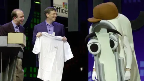 Getty Images Bill Gates smiles holding a white t-shirt next to an actor wearing a giant Clippy foam suit