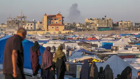 EPA Smoke rises in the distance while people stand in front of a tent community in Rafah on 7 May