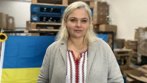 Nicola Haseler/BBC Yuliya Halahan standing in a warehouse in front of a Ukraine flag