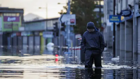 PA Media A man in waterproofs walks through knee-high floodwater in Downpatrick