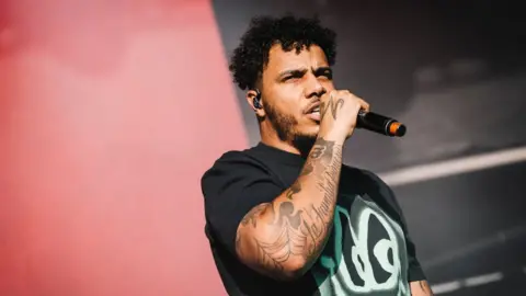 Getty Images AJ Tracey performing at Laneway festival, he is wearing a black t-shirt and holding a microphone