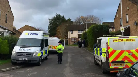 BBC Image of Wedmore Close in Kingswood. Police vans can be seen either side of the road, and a female officer is standing next to a van. Houses can be seen either side of the road and in the distance.