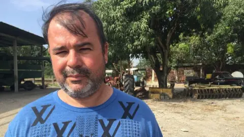 Ronald Lakra, a man with thinning hair and a greying beard, stands in his farm.