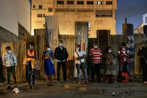 Yasuyoshi Chiba / AFP Commuters wait for a bus ahead of the night time curfew in Nairobi, on 27 March 2021
