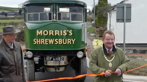 Vince Hunt and Michael Limbrey cutting a ribbon in front of waiting vintage vehicles