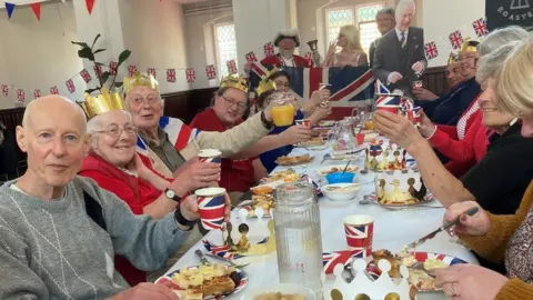 BBC Long table with people raising a union jack cup as they tuck into a coronation lunch laid on by the Storehouse foodbank in Trowbridge