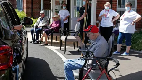 Getty Images Staff look on as a visitor uses her phone to take a picture of her grandad during a drive-through visit at Gracewell, a residential care home in Adderbury near Banbury, west of London on May 28